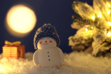 Photo of Cute decorative snowman, gift box and Christmas tree on artificial snow against dark background