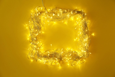 Photo of Frame made of glowing Christmas lights on yellow background, top view. Space for text