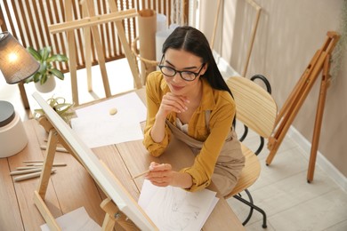 Young woman drawing on easel with pencil at table indoors, above view