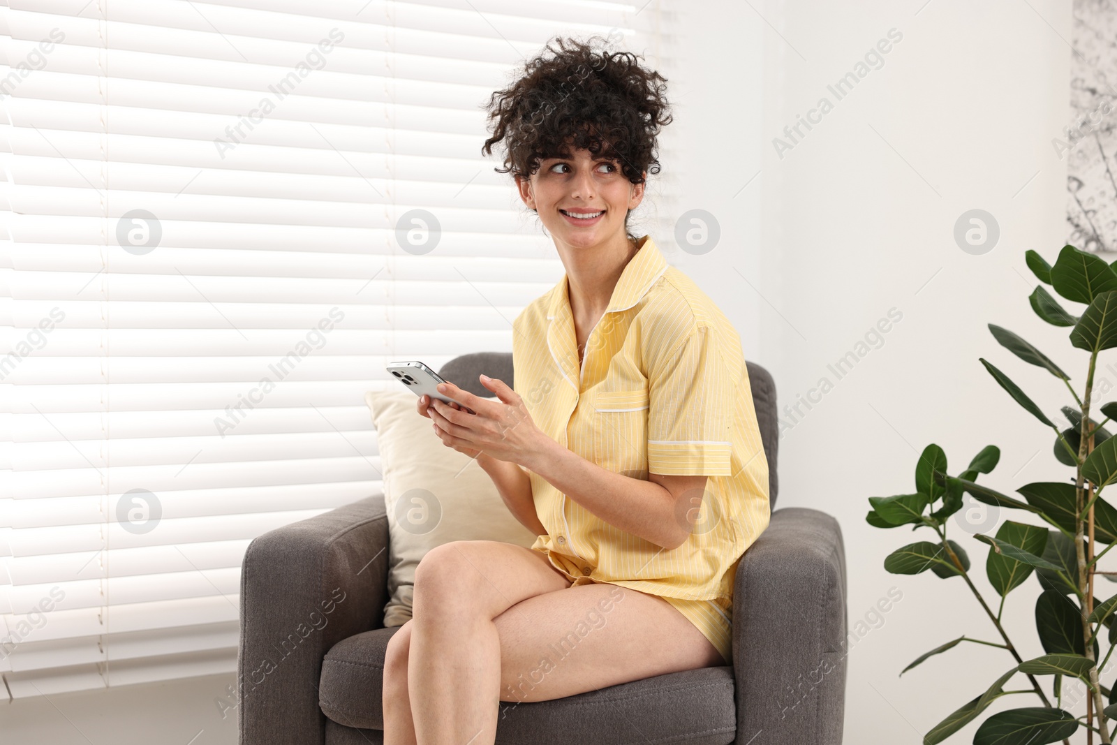 Photo of Beautiful young woman in stylish pyjama with smartphone on armchair at home