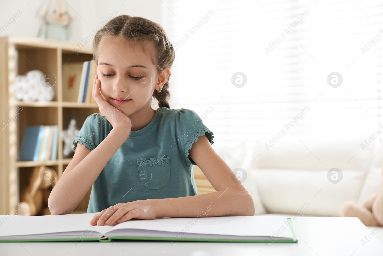 Photo of Cute little girl reading book at desk in room. Space for text