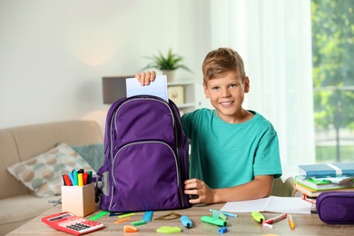 Photo of Cute boy putting school stationery into backpack at table indoors