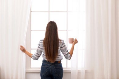 Photo of Young woman with cup of coffee opening window curtains at home