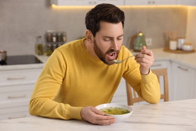 Photo of Man eating delicious chicken soup at light marble table in kitchen