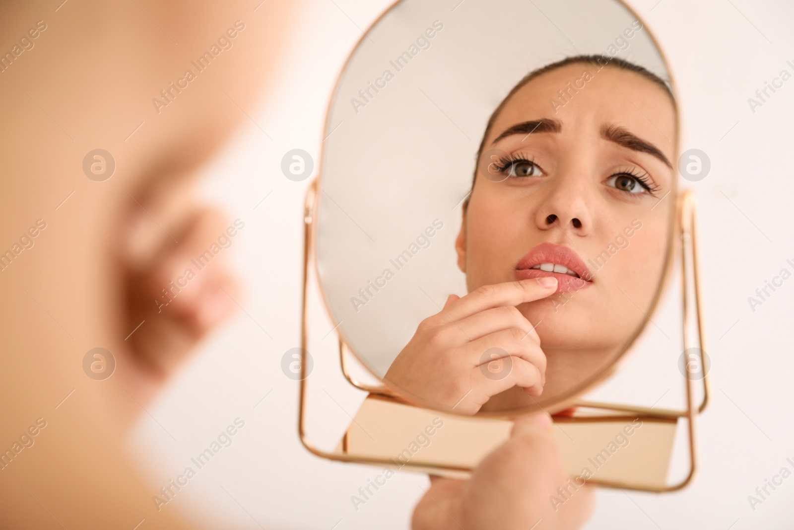 Photo of Emotional woman with herpes touching lips in front of mirror against light background