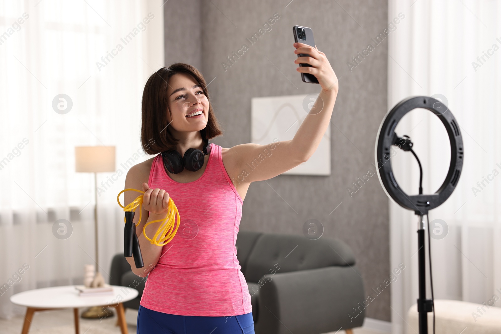 Photo of Happy sports blogger holding skipping rope while streaming online fitness lesson with smartphone at home