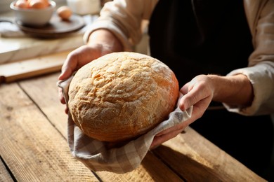 Photo of Man holding loaf of fresh bread at wooden table indoors, closeup