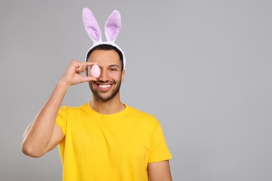 Happy African American man in bunny ears headband covering eye with Easter egg on gray background. Space for text