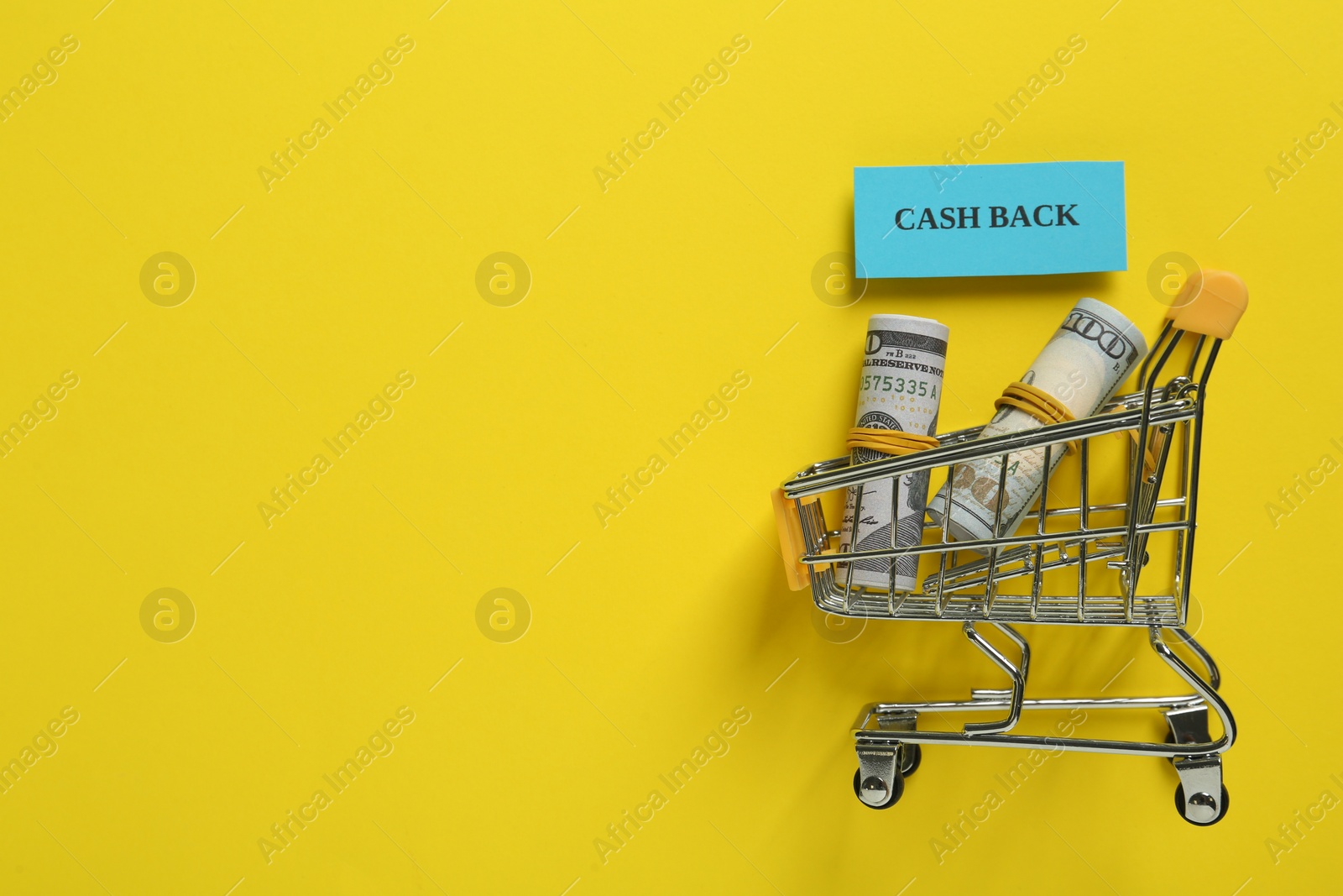 Photo of Card with word Cashback and rolled dollar banknotes in shopping cart on yellow background, flat lay. Space for text