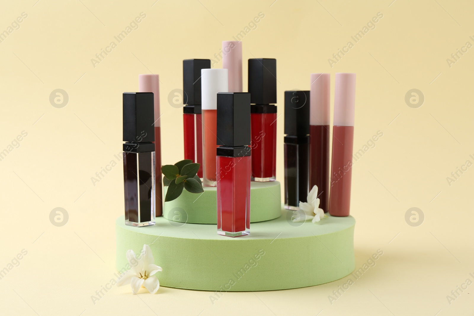 Photo of Different lip glosses, podiums and flowers on beige background