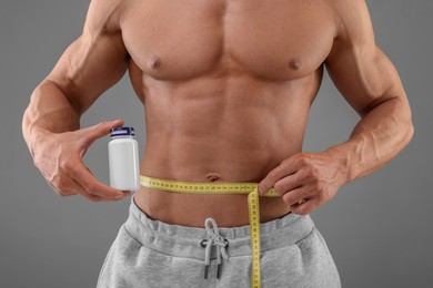 Photo of Athletic man holding bottle of supplements and measuring his waist with tape on grey background, closeup. Weight loss