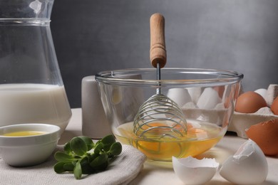 Photo of Metal whisk, raw eggs in bowl and ingredients on light table, closeup