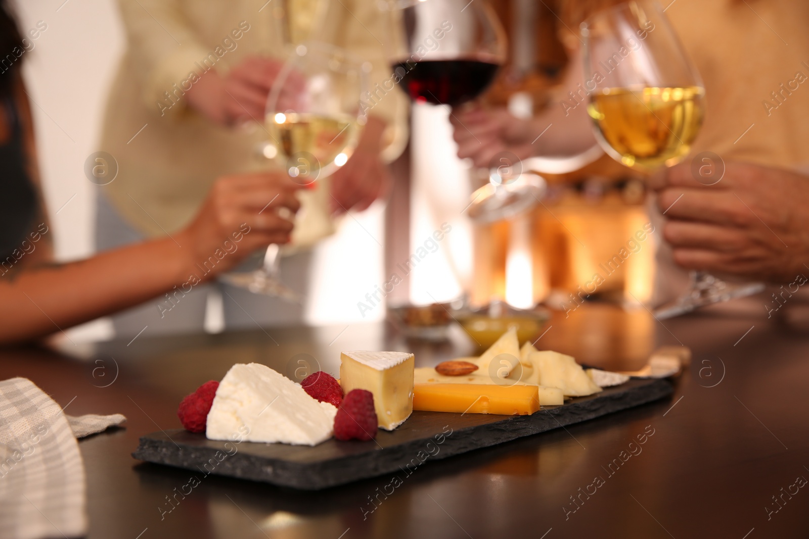 Photo of Different delicious cheeses on table and blurred friends toasting with wine on background