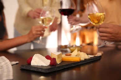 Different delicious cheeses on table and blurred friends toasting with wine on background