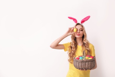 Photo of Beautiful young woman in bunny ears headband with Easter eggs on light background, space for text