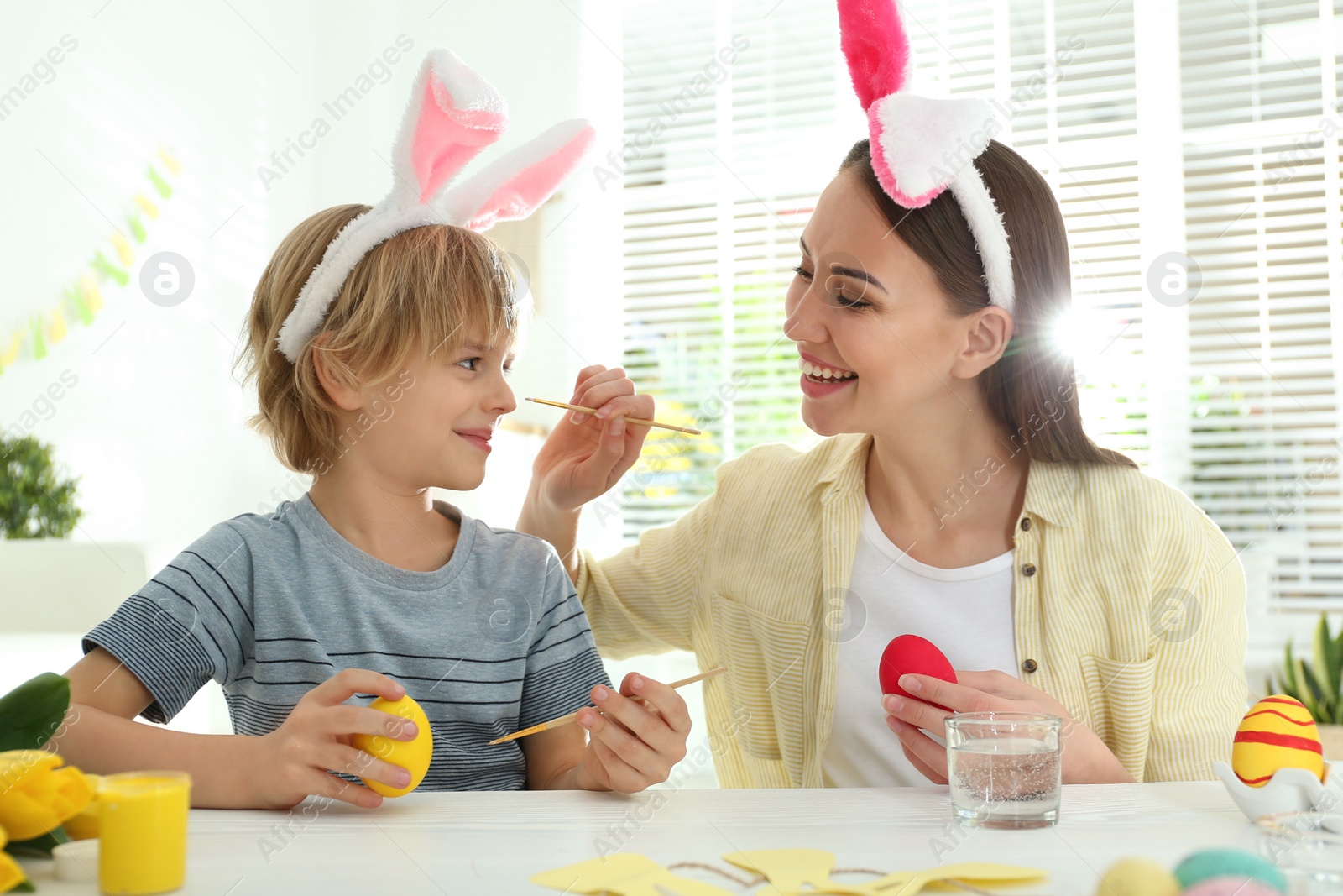 Photo of Happy mother and son with bunny ears headbands  having fun while painting Easter eggs at home