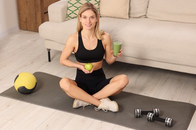 Photo of Young woman in sportswear with glass of fresh smoothie and apple at home