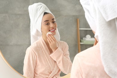 Photo of Beautiful woman in terry towel removing makeup with cotton pad near mirror indoors