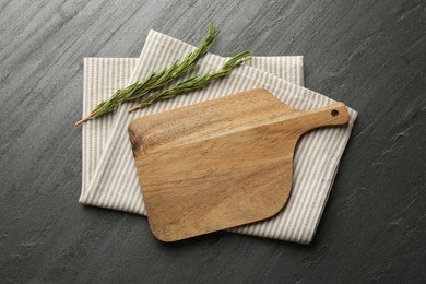 Photo of Wooden cutting board, kitchen towel and rosemary on dark grey table, top view