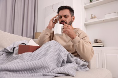 Photo of Sick man with tissue and cup of drink on sofa at home, low angle view. Cold symptoms