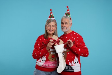 Photo of Senior couple in Christmas sweaters taking gift from stocking on light blue background