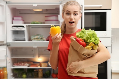 Photo of Beautiful young woman with paper bag full of food near refrigerator at home