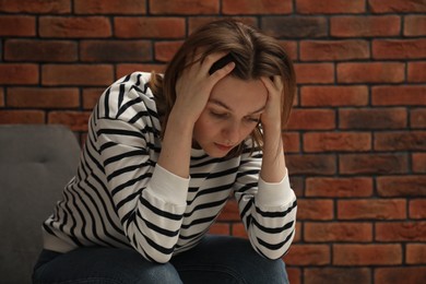 Sad young woman sitting on chair near brick wall, space for text