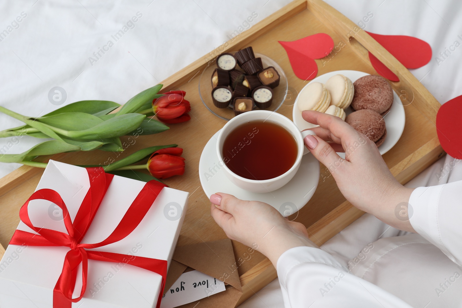 Photo of Tasty breakfast served in bed. Woman with tea, desserts, gift box and flowers at home, closeup