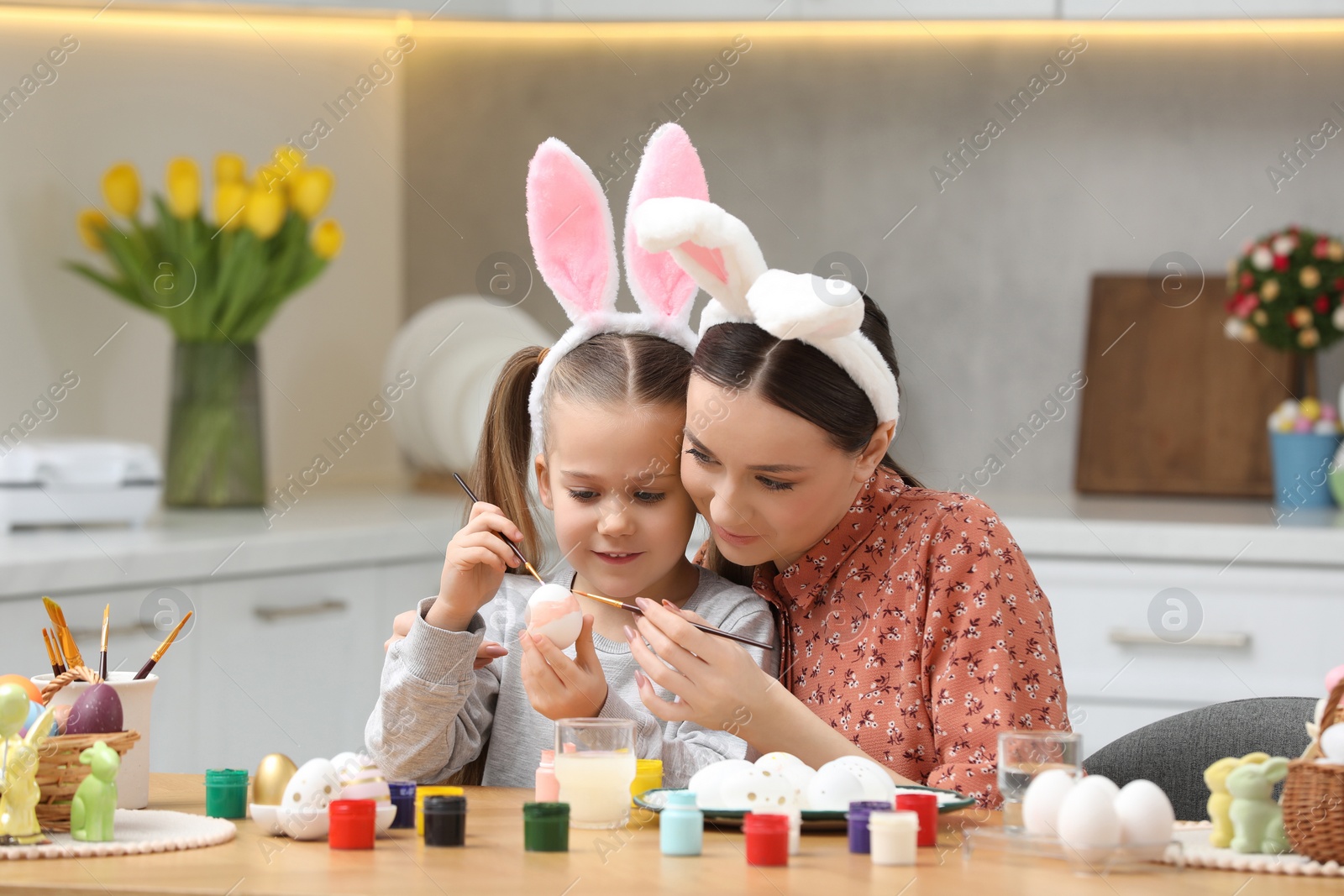 Photo of Mother and her cute daughter painting Easter eggs at table in kitchen