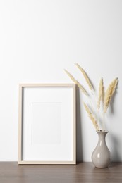 Photo of Empty photo frame and vase with dry decorative spikes on wooden table. Mockup for design