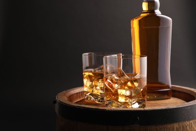 Whiskey with ice cubes in glasses and bottle on wooden barrel against black background, space for text