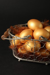 Shiny golden eggs with feathers in metal basket on black background