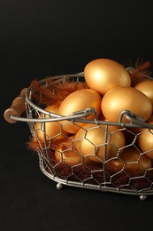 Photo of Shiny golden eggs with feathers in metal basket on black background