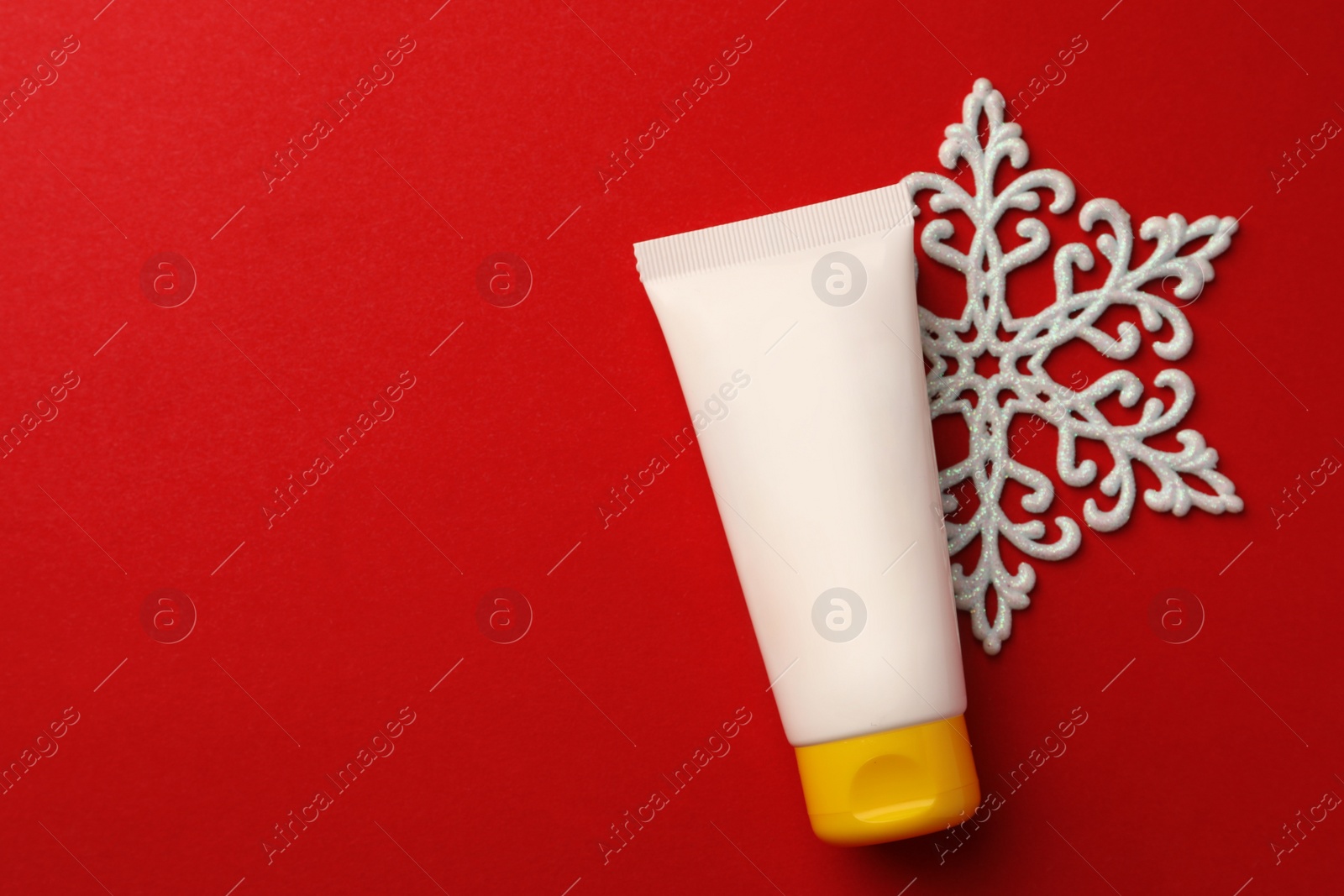 Photo of Tube of hand cream and snowflake on red background, flat lay with space for text. Winter skin care