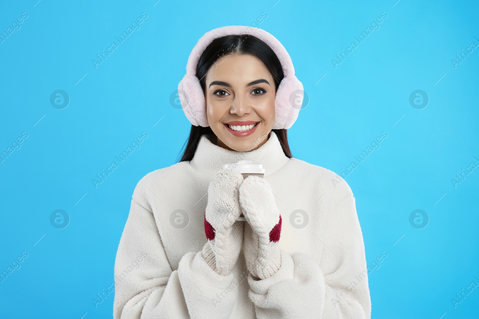 Photo of Beautiful young woman in earmuffs with cup of drink on light blue background