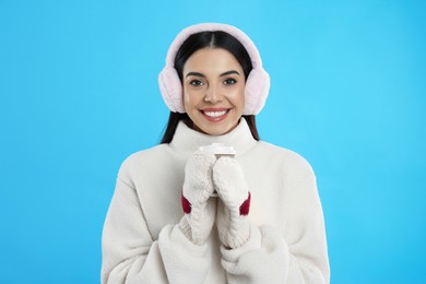 Beautiful young woman in earmuffs with cup of drink on light blue background