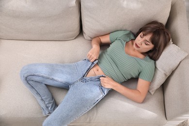 Young woman struggling to squeeze into tight jeans while lying on sofa, above view