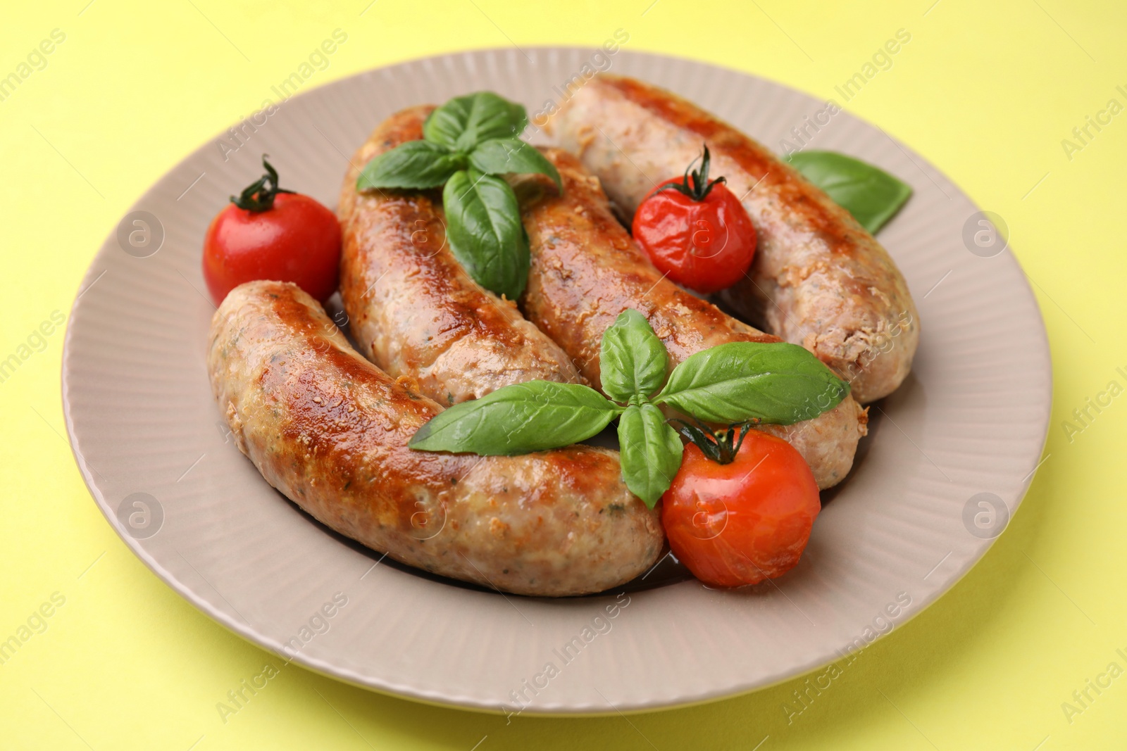 Photo of Plate with tasty homemade sausages, basil leaves and tomatoes on yellow table, closeup