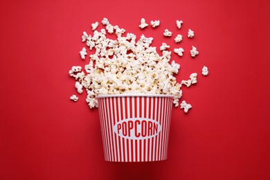 Photo of Overturned paper bucket with delicious popcorn on red background, flat lay