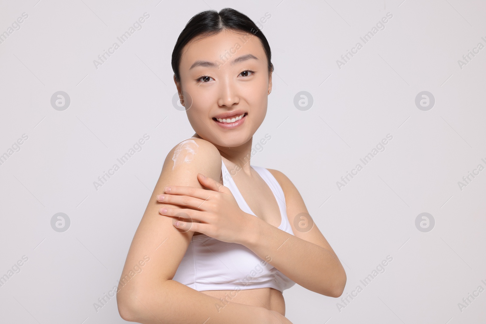 Photo of Beautiful young Asian woman applying body cream onto shoulder on light grey background
