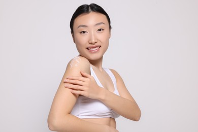 Photo of Beautiful young Asian woman applying body cream onto shoulder on light grey background