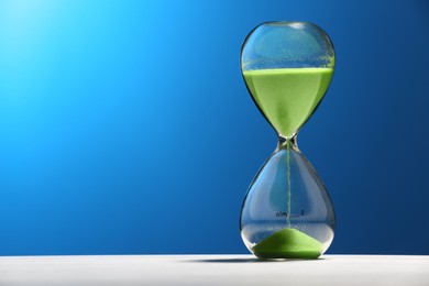 Photo of Hourglass with light green flowing sand on white table against blue background. Space for text