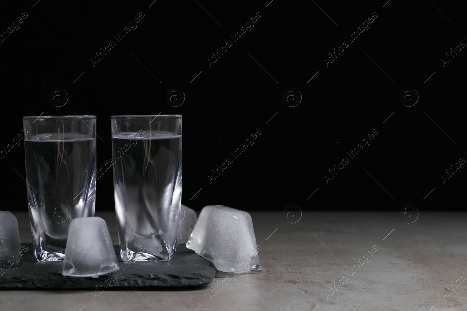 Photo of Shot glasses with vodka and ice cubes on grey tale against black background, space for text