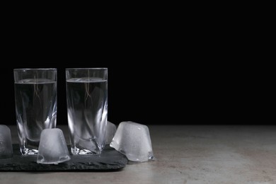 Photo of Shot glasses with vodka and ice cubes on grey tale against black background, space for text