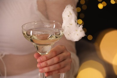 Woman holding glass of cocktail decorated with tasty cotton candy on black background with blurred lights, closeup