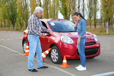 Photo of Indignant senior instructor and young woman near car outdoors. Passing driving license exam