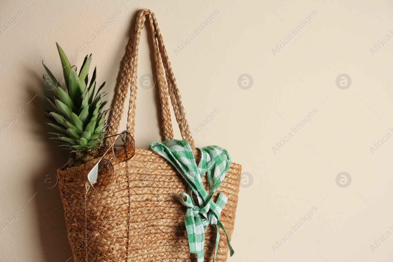 Photo of Elegant woman's straw bag with pineapple and accessories hanging on beige background. Space for text