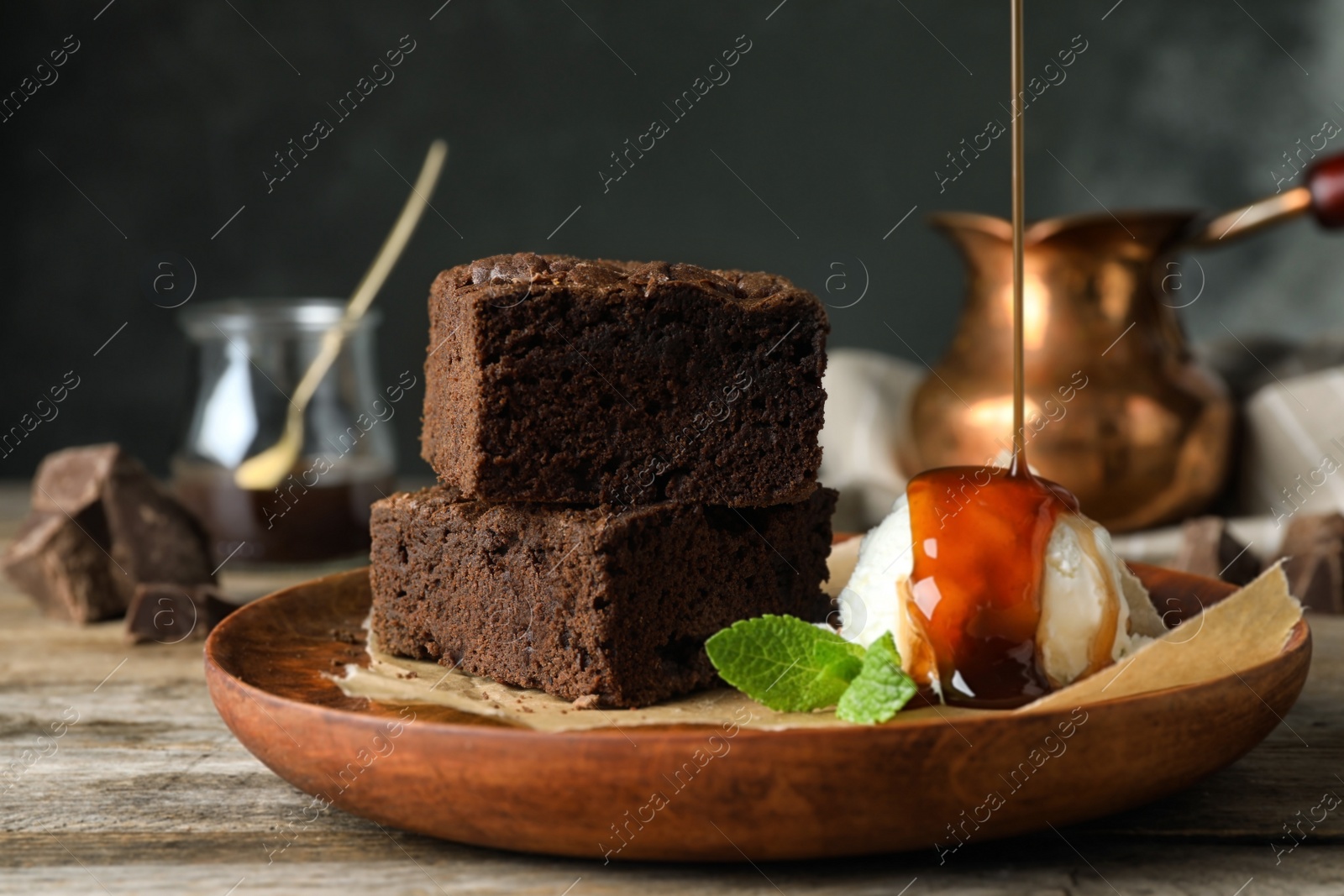 Photo of Pouring sauce onto ice-cream with fresh brownies in wooden plate on table. Delicious chocolate pie