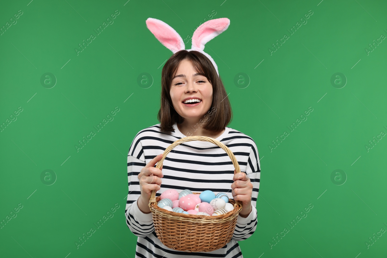 Photo of Easter celebration. Happy woman with bunny ears and wicker basket full of painted eggs on green background