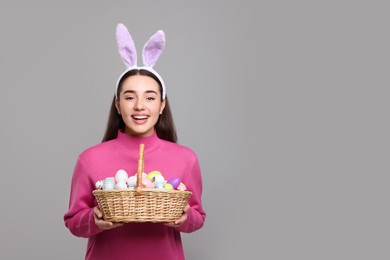 Photo of Happy woman in bunny ears headband holding wicker basket of painted Easter eggs on grey background. Space for text